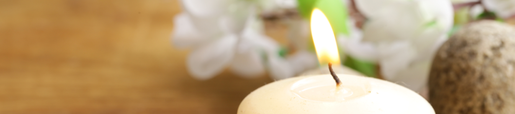 A lit candle resting on a tray with pebbles, similar to a spa. In the background, white flowers rest on a table.