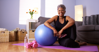 African American woman in workout clothing sitting on yoga mat next to a yoga ball and weights