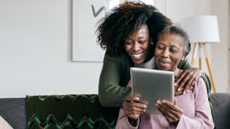 An older Black woman in a pink shirt sits on a couch holding a tablet, with a younger Black woman hugging her shoulders from behind. They smile at the screen together