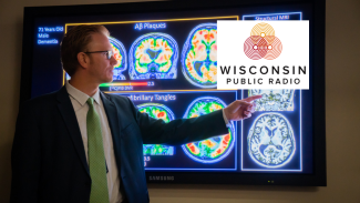 Dr. Sterling Johnson in front of a screen with multiple PET scans and MRI scans. He points on the right side of the screen, where the Wisconsin Public Radio logo has been edited on top of the screen