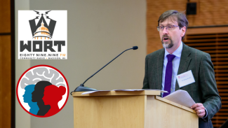 Art Walaszek, MD, stands behind a podium to talk to a crowd. The WORT-FM logo and Fall Community Conversation logo appear to the left of the podium