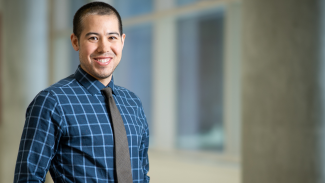 Photo of Dr. Nathaniel Chin in a hallway with windows. He's wearing a blue plaid shirt and grey tie.