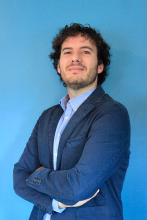 man with dark, curly hair wearing a blue suit jacket standing in front of a light blue wall