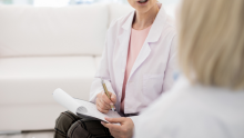 A doctor, whose face isn't seen, holding a notepad and pen as they talk with a patient