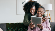 An older Black woman in a pink shirt sits on a couch holding a tablet, with a younger Black woman hugging her shoulders from behind. They smile at the screen together