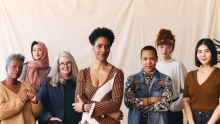A group of women, of various races, ethnicities, and ages, stand together in front of an off-white background