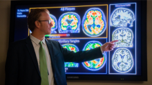 Sterling Johnson, PhD, stands in front of a monitor of several PET and MRI scans. He points at an MRI in the middle of a column of three MRIs.