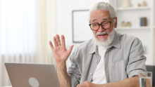 An older adult sits at a desk using a laptop and raises a hand in a greeting during a video call in a bright, home office setting.