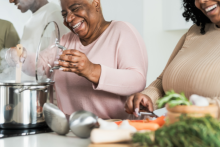 People cooking together in a kitchen, with one person stirring a pot on the stove while another chops vegetables on a cutting board.
