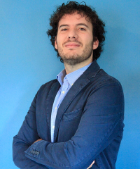 man with dark, curly hair wearing a blue suit jacket standing in front of a light blue wall